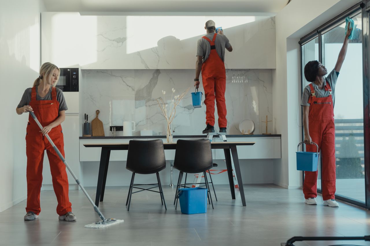 Groupe de nettoyeurs en uniformes rouges passant la serpillière et essuyant une vitre dans une salle à manger moderne.